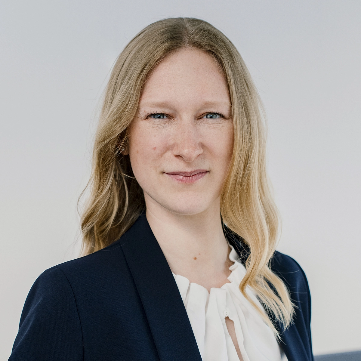 Eine junge Frau mit langen, blonden Haaren trägt einen schwarzen Blazer und eine weiße Bluse. Sie schaut ernst in die Kamera und steht vor einem neutralen Hintergrund.