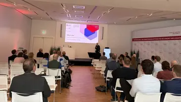 Ein Vortragender steht vor einem Publikum in einem modernen Raum. Auf einer Leinwand wird eine Präsentation angezeigt, die das Thema „MYCode“ behandelt. Die Zuschauer sitzen in weißen Stühlen und sind auf den Referenten fokussiert.
