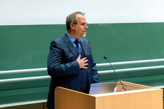 Ein Mann in einem Anzug steht an einem Rednerpult und spricht. Er hat eine Hand auf seinem Herzen gelegt und schaut aufmerksam in die Zuhörerschaft. Im Hintergrund ist eine grüne Tafel zu sehen.