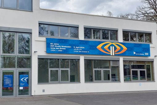 Blick auf einen großen Screen an einer Gebäudefassade, darauf Werbung für die Jubiläumsveranstaltung 60 Jahre ZDF Unter den Eichen