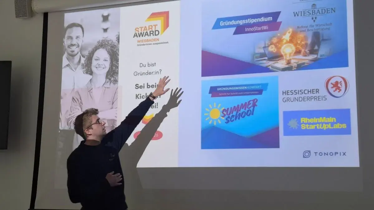 Ein Mann steht an einer Präsentationswand und zeigt auf verschiedene Logos und Textinformationen zu Gründerinitiativen wie „START AWARD“, „Gründungstipendium“ und „Summer School“. Der Text ermutigt Gründerinnen zur Teilnahme an den Programmen und Veranstaltungen.