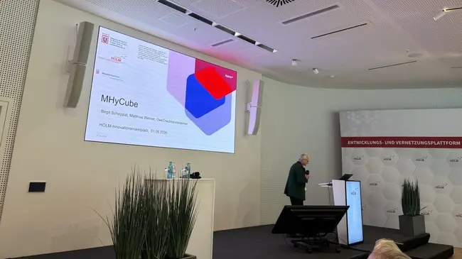 Ein Referent steht vor einer Präsentation mit dem Titel "MHyCube". Auf der Folie sind grafische Elemente in Rot und Blau sowie Logos zu sehen. Im Hintergrund befindet sich eine moderne Raumgestaltung mit Pflanzen und einer hellen Wand.
