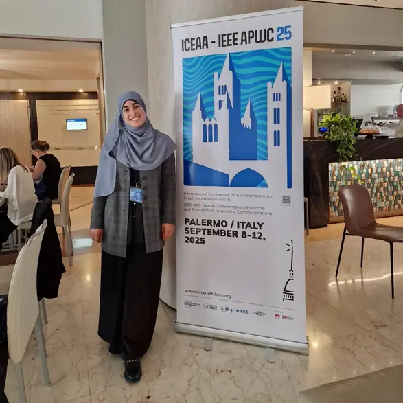 Eine junge Frau mit einem Kopftuch steht lächelnd neben einem Banner, das die Veranstaltung "ICEAA - IEEE APWC 25" ankündigt. Das Banner zeigt stilisierte Gebäude und Informationen zur Konferenz, die im September 2025 in Palermo, Italien, stattfindet.