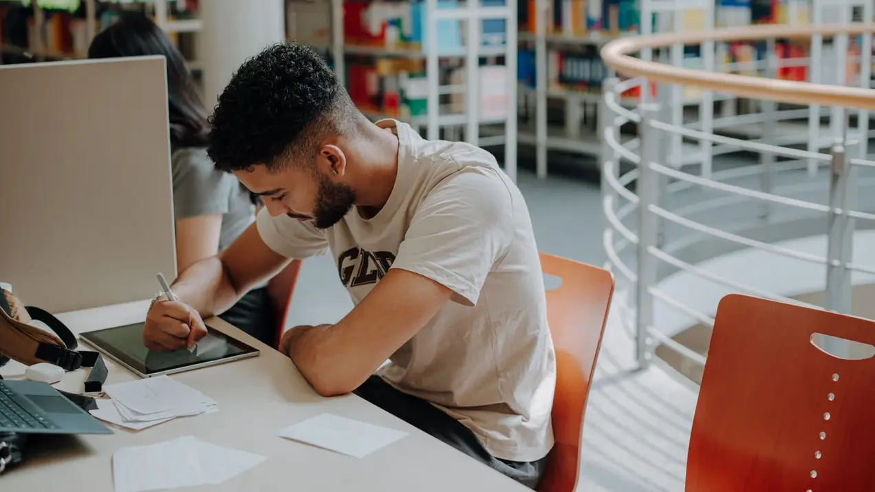 Ein Studierender sitzt an einem Einzelarbeitsplatz in der Bibliothek und schreibt etwas in sein Tablett