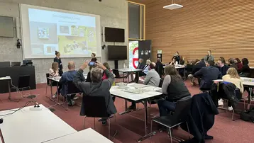Eine Gruppe von Teilnehmenden der UNESCO Jahrestagung sitzt in einem Seminarraum mit Tischen und Stühlen. Ein Bildschirm im Hintergrund zeigt eine Präsentation zu interdisziplinären Spielen. Zwei Personen stehen vorne und sprechen zu den Anwesenden, die aufmerksam zuhören.