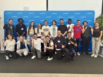 Eine Gruppe von 15 Menschen steht lächelnd vor einer blauen Wand mit dem Logo von Boeing. Alle heben die Hände und begrüßen freundlich. Die Gruppe besteht aus verschiedenen Personen mit unterschiedlichen Hintergründen und Stilen.