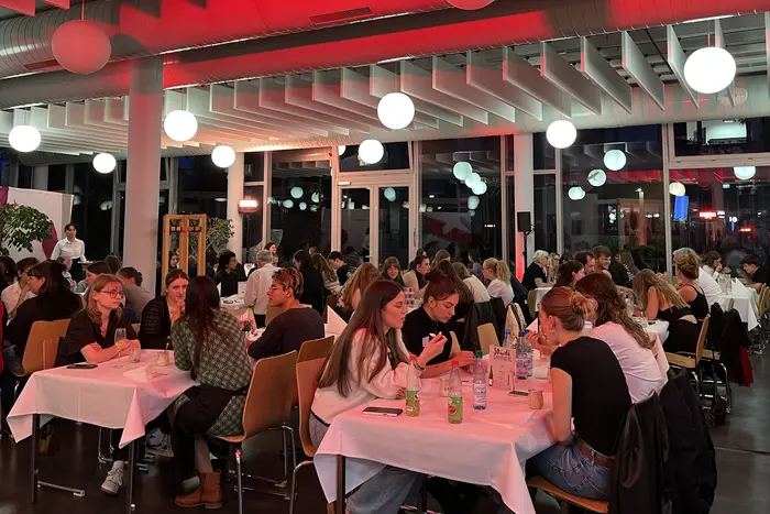 Die Gäste sitzen an Tischen und unterhalten sich, während im Hintergrund sanftes, rotes Licht von der Decke strahlt. Helle Lampen hängen von der Decke und schaffen eine einladende Atmosphäre.
