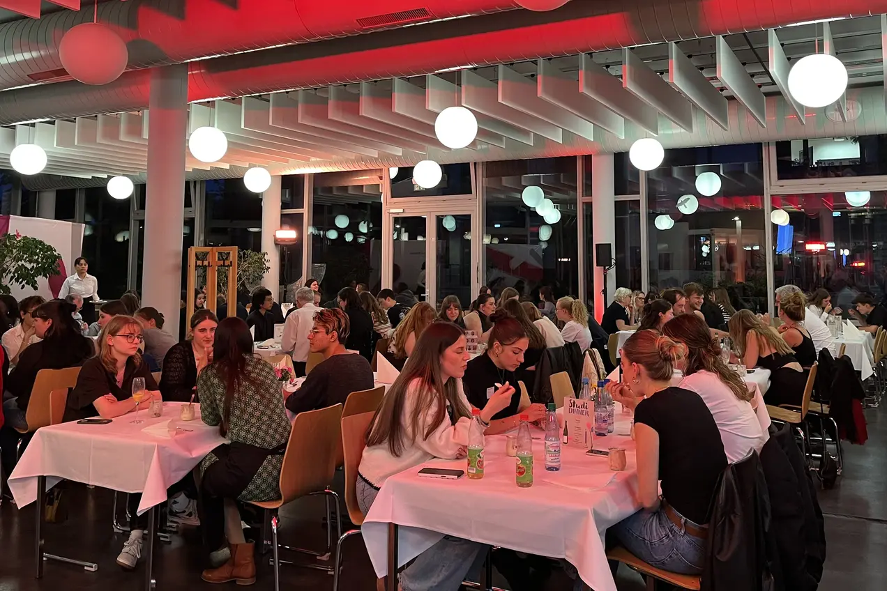Die Gäste sitzen an Tischen und unterhalten sich, während im Hintergrund sanftes, rotes Licht von der Decke strahlt. Helle Lampen hängen von der Decke und schaffen eine einladende Atmosphäre.