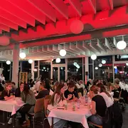 Die Gäste sitzen an Tischen und unterhalten sich, während im Hintergrund sanftes, rotes Licht von der Decke strahlt. Helle Lampen hängen von der Decke und schaffen eine einladende Atmosphäre.