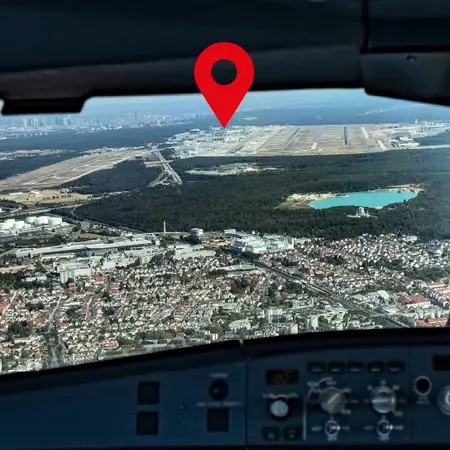 Blick aus dem Cockpit eines Flugzeugs über eine Landschaft mit einem markierten Punkt. Der Punkt deutet auf einen Flughafen hin, während sich darunter ein See und städtische Gebiete befinden.