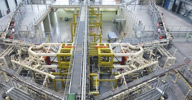 Eine hochmoderne Maschinenanlage in einem Industriegebäude. Im Zentrum befinden sich verschiedene mechanische Komponenten, die durch Förderbänder verbunden sind. Die Einrichtung scheint für Automatisierungs- oder Produktionsprozesse konzipiert zu sein, mit gesicherten Geländern und einem großen, offenen Raum im Hintergrund.
