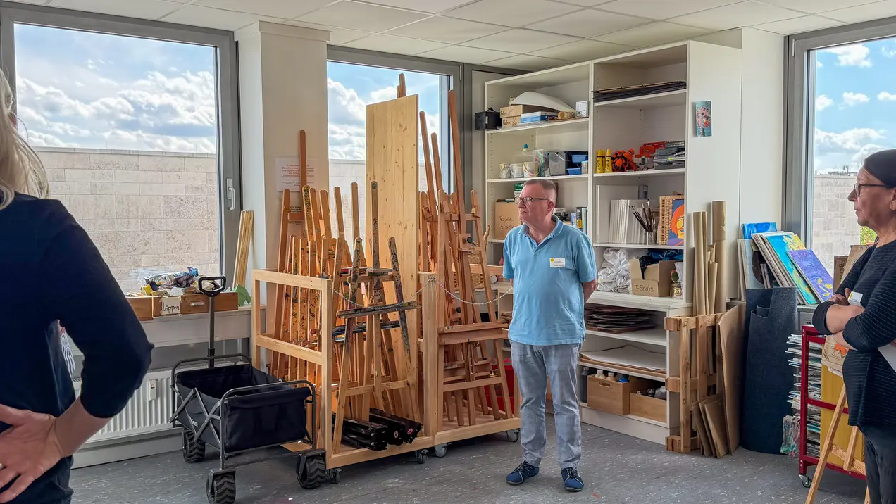 In einem Atelier stehen zwei Personen, eine links und eine rechts, während ein Mann in der Mitte spricht. Im Hintergrund sieht man viele Staffeleien und Regale mit Kunstmaterialien. Das Licht strömt durch große Fenster, die auf einen bewölkten Himmel blicken.