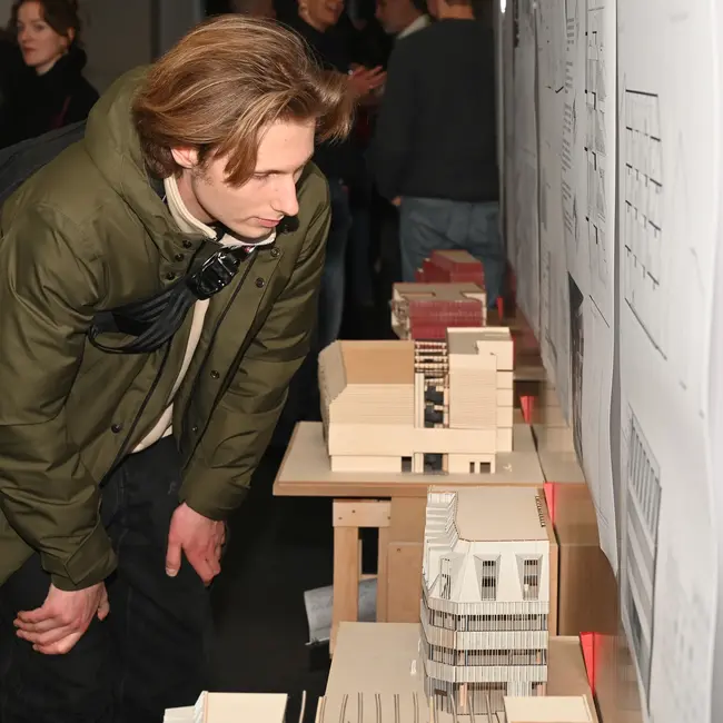 Ein Student beugt sich über einen Tisch mit Architekturmodellen. Im Hintergrund sind mehrere Personen zu sehen, die an Wänden mit Plänen und Zeichnungen stehen. Es handelt sich um die Ausstellung der Bachelor- und Master-Thesen im Studiengang Architektur.