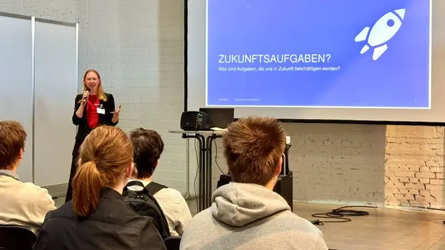 Eine Frau steht vor einer Präsentation mit dem Titel „Zukünftige Aufgaben?“ auf einem blauem Hintergrund. Sie spricht zu einem Publikum, das aus mehreren Personen in einem Seminarraum besteht. Die Präsentation und die Rednerin sind im Vordergrund, während die Zuhörer im Hintergrund sitzen.
