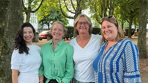 Vier Frauen stehen lächelnd in einem Park unter Bäumen. Sie tragen unterschiedliche, sommerliche Kleidung und stehen eng beieinander. Der Hintergrund zeigt einen klaren Himmel und weiteren Baumbewuchs, was eine freundliche und einladende Atmosphäre vermittelt.