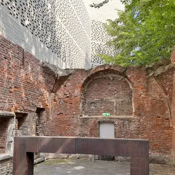 The picture shows the historic church area of the Kolumba Museum in Cologne, with a simple, contemporary altar in the centre. In the background, extending from the brick wall, is the Kolumba Museum building, constructed from narrow white bricks.