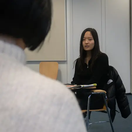 Eine Frau mit langen, dunklen Haaren sitzt auf einem Stuhl und schaut aufmerksam. Sie trägt ein schwarzes Oberteil. Im Vordergrund ist der Hinterkopf einer anderen Person zu sehen, die ebenfalls auf einem Stuhl sitzt. Der Raum hat eine helle, neutrale Farbgebung.