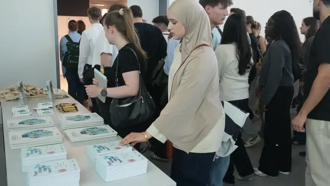 Eine Gruppe von Menschen steht an einem Tisch mit mehreren Stapeln von Büchern. Eine Frau mit einem Kopftuch betrachtet die Bücher und greift nach einem Exemplar. Im Hintergrund sind weitere Personen zu sehen, die sich ebenfalls für die Bücher interessieren.