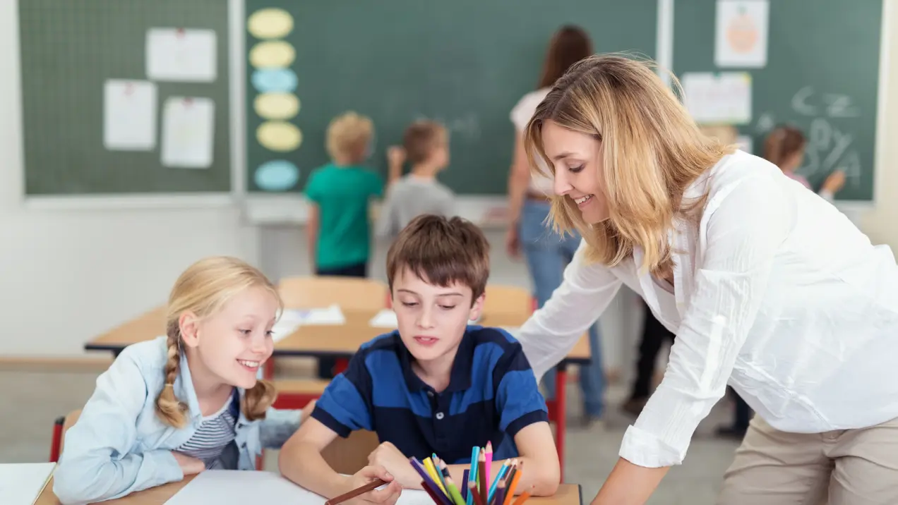Eine Lehrerin unterstützt zwei Kinder beim Lernen an einem Tisch. Die Kinder schauen auf ein Heft und haben Buntstifte vor sich. Im Hintergrund sind eine weitere Lehrerin und weitere Schüler:innen zu sehen, die vor einer Tafel stehen.