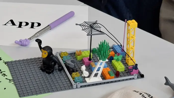 Eine LEGO-Szene zeigt eine Figur in schwarzem Anzug, die einen Arm erhoben hat. Um sie herum sind bunte Bausteine, eine grüne Pflanze und ein Netz. Im Hintergrund steht ein gelbes Gerüst. Auf dem Tisch liegt ein Blatt mit der Aufschrift "App".