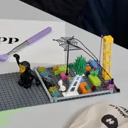 Eine LEGO-Szene zeigt eine Figur in schwarzem Anzug, die einen Arm erhoben hat. Um sie herum sind bunte Bausteine, eine grüne Pflanze und ein Netz. Im Hintergrund steht ein gelbes Gerüst. Auf dem Tisch liegt ein Blatt mit der Aufschrift "App".