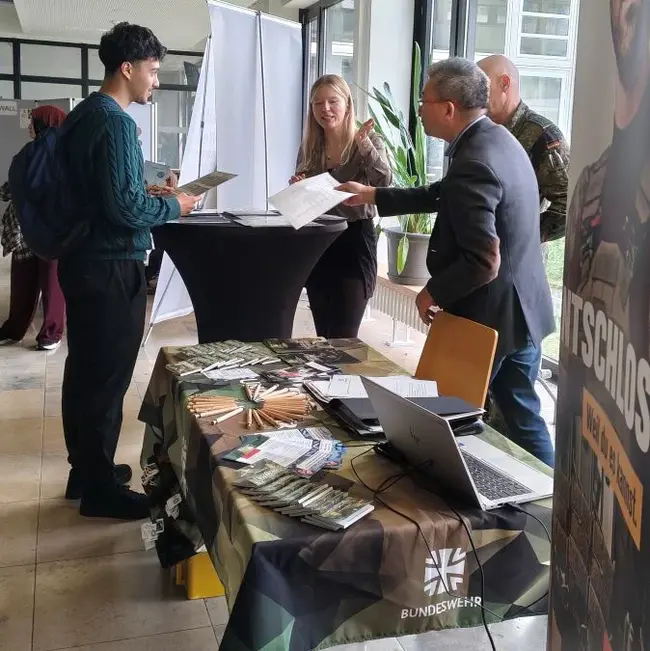 Ein Stand mit Mitarbeitern, die Informationen an Interessierte ausgeben. Eine Frau und zwei Männer stehen an einem Tisch, der mit Prospekten und Materialien dekoriert ist. Im Hintergrund sind weitere Personen und ein Banner zu sehen, der für die Bundeswehr wirbt.