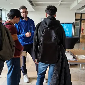 Drei Personen stehen in einem Gebäude und unterhalten sich intensiv. Eine Person trägt einen blauen Hoodie, während die anderen in lässiger Kleidung sind. Im Hintergrund sind weitere Sitzgelegenheiten und ein Bildschirm sichtbar.