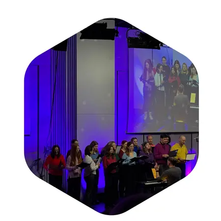 Eine Gruppe von zehn Personen steht auf der Bühne und singt. Sie tragen bunte Oberteile in verschiedenen Farben. Im Hintergrund ist ein Klavier sichtbar, und eine Projektionsleinwand zeigt sie beim Singen. Die Atmosphäre wirkt festlich und einladend.
