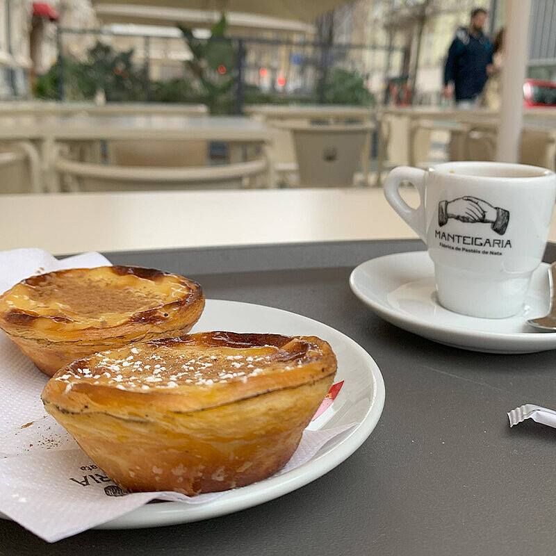 Auf einem Teller liegt Pasteis de Nata, ein Gebäck aus Lissabon. Daneben steht eine Tasse.
