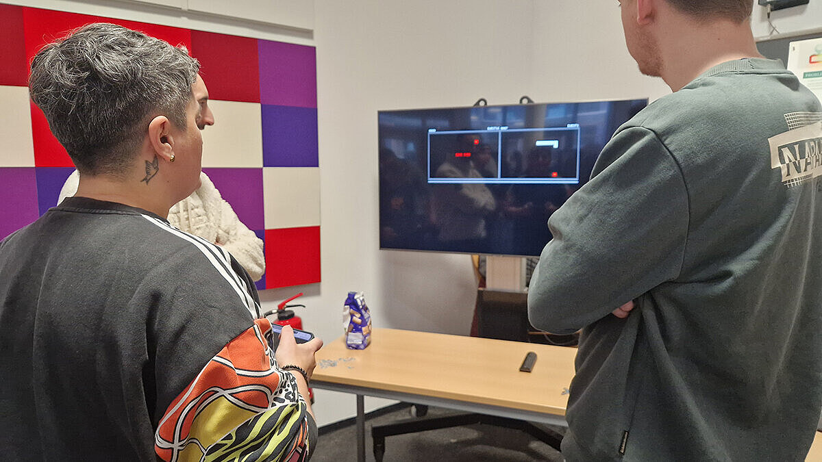 Drei Studierende stehen vor einem Bildschirm, eine Studierende hält ein Smartphone in der Hand, mit dem sie das Spiel steuert 