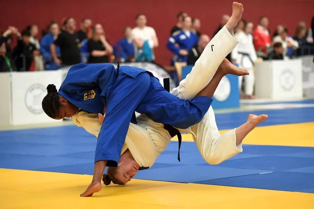 Zwei Judoka in einem Wettkampf. Die Kämpferin in blauen Judogien hebt die Gegnerin in einem Wurf. Beide Athletinnen sind auf einer blauen Judomatte, während Zuschauer:innen im Hintergrund ihre Aufmerksamkeit auf das Geschehen richten.