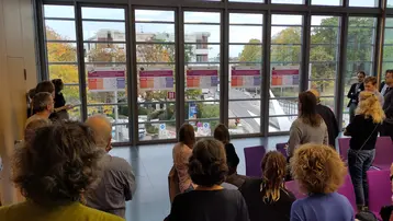 Eine Gruppe von Menschen steht in einem Raum mit großen Fenstern und diskutiert vor Präsentationen, die an der Wand hängen. Draußen sind Bäume zu sehen, die in herbstlichen Farben leuchten. Die Szene vermittelt eine Atmosphäre von Austausch und Zusammenarbeit.