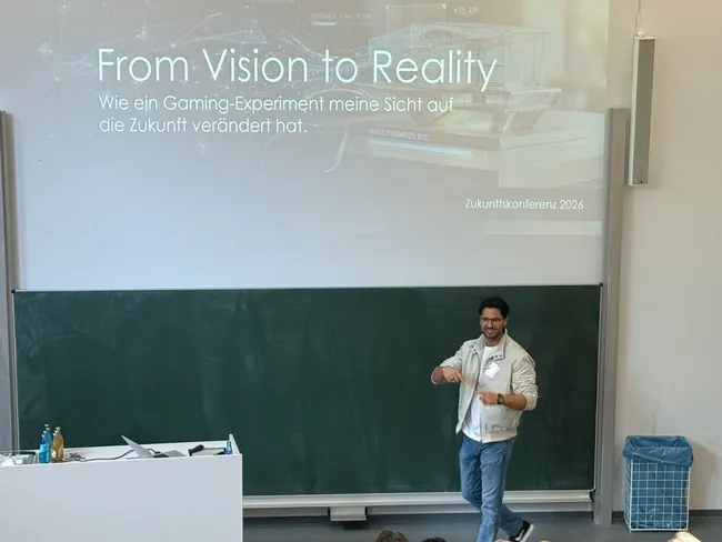 Ein Referent steht auf einer Bühne und spricht zu einem Publikum. Im Hintergrund ist eine Präsentation mit dem Titel „From Vision to Reality“ zu sehen, die sich mit einem Gaming-Experiment und dessen Einfluss auf die Zukunft beschäftigt.