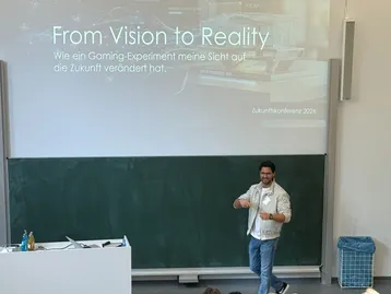 Ein Referent steht auf einer Bühne und spricht zu einem Publikum. Im Hintergrund ist eine Präsentation mit dem Titel „From Vision to Reality“ zu sehen, die sich mit einem Gaming-Experiment und dessen Einfluss auf die Zukunft beschäftigt.