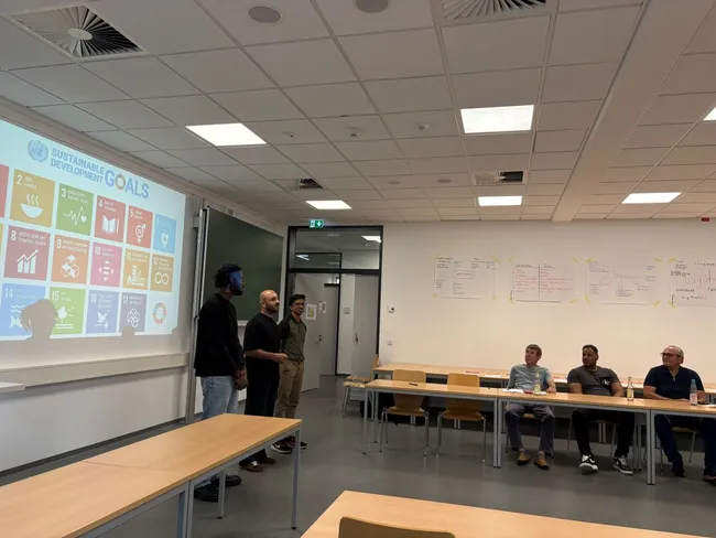 Eine Präsentation über die Nachhaltigkeitsziele der Vereinten Nationen. Im Vordergrund stehen zwei Personen, die sprechen, während im Hintergrund eine Gruppe von vier Menschen sitzt. An einer Wand sind Notizen sichtbar, die den Kontext des Themas unterstützen.