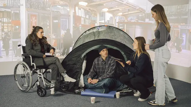 Vier Personen vor einer Leinwand welche einen Bahnhof zeigt. Eine Person stellt einen Obdachlosen dar, eine Person im Rollstuhl und zwei Personen, die als Sozialarbeiter:innen funigeren. Der Obdachlose sitzt auf dem Boden in einem Zelt und hört den Sozialarbeiter:innen gespannt zu, so wie die Person im Rollstuhl.