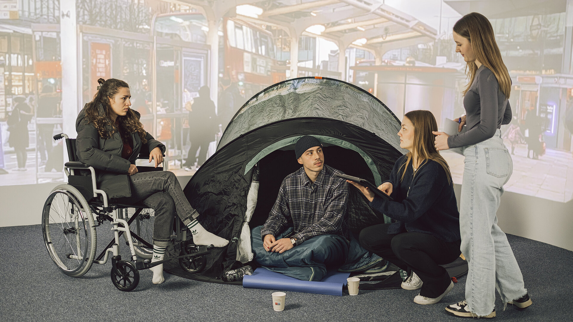 Vier Personen vor einer Leinwand welche einen Bahnhof zeigt. Eine Person stellt einen Obdachlosen dar, eine Person im Rollstuhl und zwei Personen, die als Sozialarbeiter:innen funigeren. Der Obdachlose sitzt auf dem Boden in einem Zelt und hört den Sozialarbeiter:innen gespannt zu, so wie die Person im Rollstuhl.