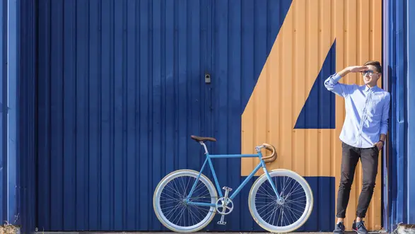 Ein junger Mann in einem hellblauen Hemd steht neben einem weißen Fahrrad vor einer blauen Wand mit einer großen orangefarbenen Zahl 4.