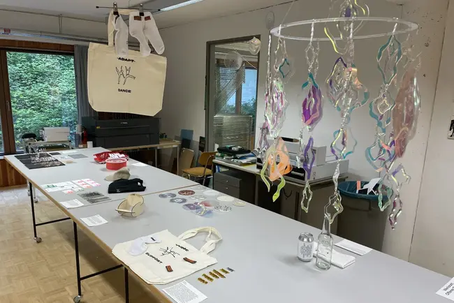 Auf einem Tisch sind verschiedene Gegenstände ausgestellt, darunter eine Tragetasche, Socken, Aufkleber, CDs und Glasbehälter. Eine farbige Mobileskulptur hängt von der Decke. Der Raum hat große Fenster, durch die Licht strömt, und eine ruhige, kreative Atmosphäre.