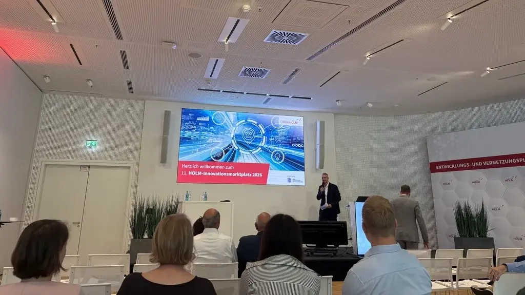 Ein Mann steht vor einem Bildschirm in einem modernen Konferenzraum. Der Bildschirm zeigt Grafiken und Text zur Veranstaltung, während das Publikum ihm zuhört. Die Atmosphäre wirkt professionell und informativ.