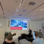 Ein Mann steht vor einem Bildschirm in einem modernen Konferenzraum. Der Bildschirm zeigt Grafiken und Text zur Veranstaltung, während das Publikum ihm zuhört. Die Atmosphäre wirkt professionell und informativ.