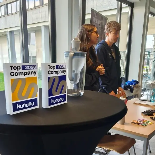 Zwei Auszeichnungen „Top Company 2023“ von kununu stehen auf einem Tisch, daneben ein Wasserglas. Im Hintergrund sind zwei Personen zu sehen, die miteinander sprechen. Die Szene vermittelt eine positive Atmosphäre, vermutlich bei einer Unternehmensveranstaltung.