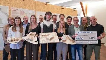 Die Abbildung zeigt eine Gruppe von Studierenden und Betreuenden. Einige halten Architekturmodelle von Gebäuden, eine Person hält einen Scheck über 2.000 Euro in den Händen. Im Hintergrund sind die Poster der Studierenden mit ihren Entwürfen zu sehen.
