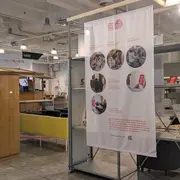 Das Bild zeigt eine Ausstellung in einem modernen Raum. Im Vordergrund befindet sich ein großer, informierender Banner mit mehreren Bildern, die Aktivitäten und Gemeinschaftsprojekte darstellen. Links steht ein holzfarbenes Möbelstück, während rechts gemütliche Sitzecke zu sehen ist.