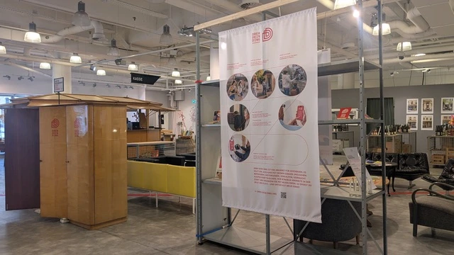 Das Bild zeigt eine Ausstellung in einem modernen Raum. Im Vordergrund befindet sich ein großer, informierender Banner mit mehreren Bildern, die Aktivitäten und Gemeinschaftsprojekte darstellen. Links steht ein holzfarbenes Möbelstück, während rechts gemütliche Sitzecke zu sehen ist.