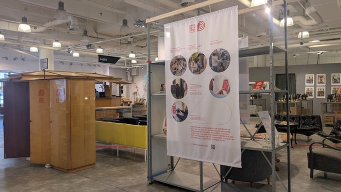 Das Bild zeigt eine Ausstellung in einem modernen Raum. Im Vordergrund befindet sich ein großer, informierender Banner mit mehreren Bildern, die Aktivitäten und Gemeinschaftsprojekte darstellen. Links steht ein holzfarbenes Möbelstück, während rechts gemütliche Sitzecke zu sehen ist.