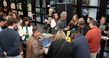 Eine Gruppe von Menschen steht um einen Tisch in einem Buchladen oder einer Bibliothek. Einige Personen unterhalten sich, während andere Bücher begutachten. Die Atmosphäre ist lebhaft und gesellig, was auf ein Event oder eine Buchpräsentation hindeutet.