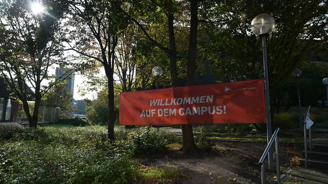 Ein rotes Banner mit der Aufschrift "Willkommen auf dem Campus!" hängt zwischen Bäumen. Die Umgebung zeigt eine grüne Landschaft mit Stufen und Laternen. Der Himmel ist hell, es ist Tag.