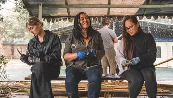 Drei Frauen sitzen auf einer Bank und arbeiten mit Werkzeugen in einem handwerklichen Umfeld. Eine Frau in der Mitte lächelt, während die anderen konzentriert an ihren Projekten arbeiten. Im Hintergrund sind verschwommen weitere Personen und eine überdachte Fläche zu sehen.