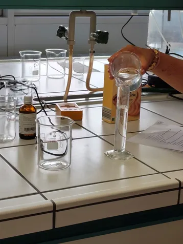 Eine Person gießt Flüssigkeit aus einem Messbecher in einen hohen Zylinder in einem Labor. Auf dem Tisch stehen weitere Gläser und eine Flasche mit einer chemischen Lösung. Das Labormaterial deutet auf eine chemische Experimentsituation hin.